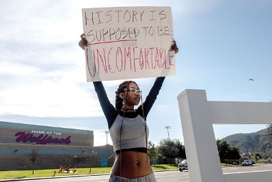 Student protester holding a sign that reads “History is supposed to be uncomfortable” outside a school district building.